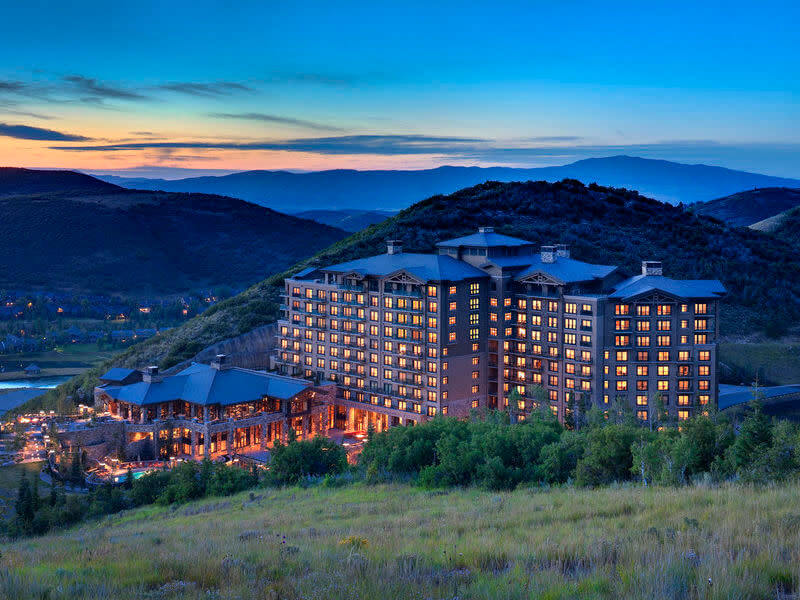 St. Regis Deer Valley hotel complex illuminated at dusk, surrounded by lush greenery and rolling hills. A serene evening sky with a gradient of blue tones stretches above, enhancing the tranquil setting.