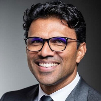 A man with short curly black hair, wearing glasses and a dark suit with a tie, smiles at the camera against a neutral gray background.