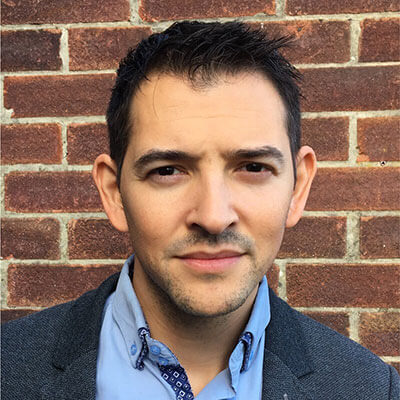 A man with short dark hair wearing a blue collared shirt, patterned tie, and gray blazer stands in front of a brick wall, looking directly at the camera with a neutral expression.