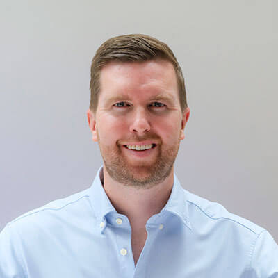A man with short brown hair and a trimmed beard smiles at the camera. He is wearing a light blue button-up shirt and stands in front of a plain, light-colored background.