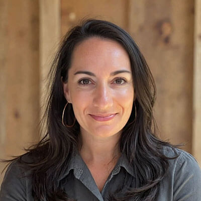 A woman with long dark hair, wearing hoop earrings and a gray collared shirt, smiles softly at the camera in front of a wooden background.