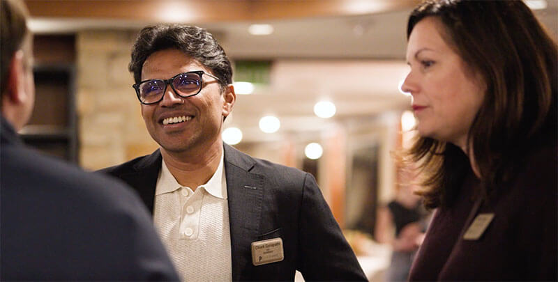 Two people wearing name tags are talking and smiling at an indoor event, with blurred background lights and another person partially visible in the foreground.