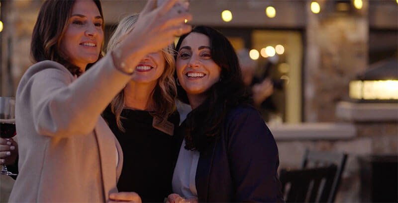 Three women smiling and taking a selfie together at an outdoor evening event, with warm string lights and a blurred background. One woman holds a glass of red wine.