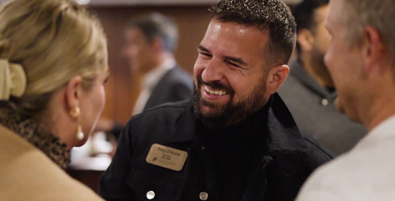 A man wearing a name tag smiles warmly while talking with two people at a social or networking event. The group appears engaged and happy, in a lively indoor setting.