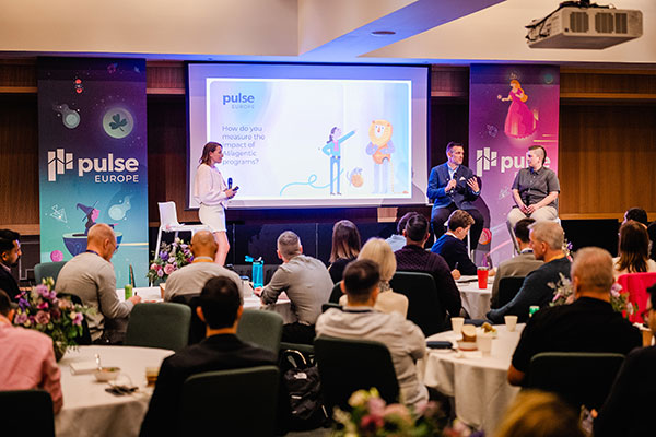 A conference scene shows a woman standing and two men seated on a stage, speaking to an audience. A screen behind them displays the gainsight pulse EUROPE logo and a slide with cartoon figures. Tables with attendees fill the room.