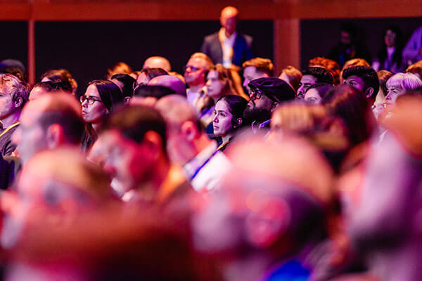 A diverse audience sits closely together in a dimly lit auditorium at Gainsight Pulse, attentively watching a presentation or speaker on stage. The lighting casts a warm, colorful glow over the crowd.