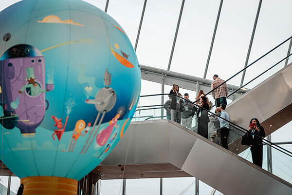 A large, colorful hot air balloon with whimsical illustrations is displayed indoors near an escalator at a Gainsight Pulse event, where several people stand and admire it inside a modern glass building.
