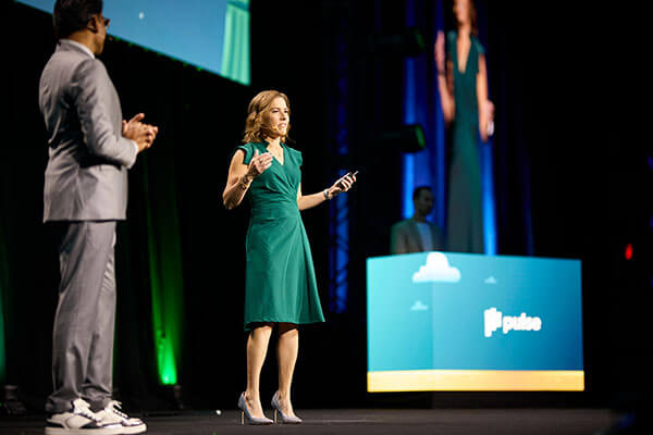 A woman in a green dress speaks on stage with a microphone, while a man in a gray suit stands nearby. Behind them, a large screen displays her image and the word 