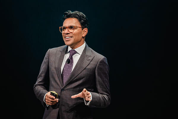 A man in a suit and tie, wearing glasses, smiles while speaking on stage at Gainsight Pulse against a dark background. He holds a small device in one hand and gestures with the other.