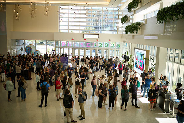 A large group of people gathers in a bright, modern convention center lobby with high ceilings and hanging plants. A colorful sign reading 