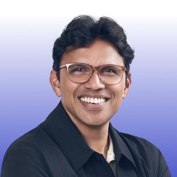 Portrait of Chuck Ganapathi, a smiling man with short, dark curly hair and glasses, wearing a black jacket over a light shirt, poses against a blue and white gradient background at Gainsight Pulse.