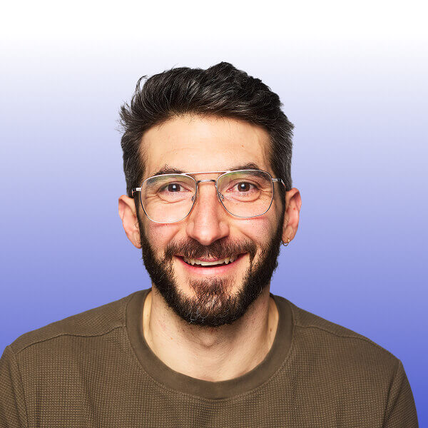Portrait of JJ Janikis, a smiling man with a beard and glasses wearing a brown sweater poses confidently in front of a gradient blue and white background, ready to speak at Gainsight Pulse.