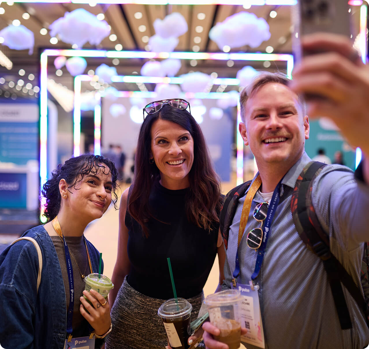 Three people smile and pose for a selfie at a colorful, well-lit Gainsight Pulse event, holding drinks and wearing lanyards. Decorative clouds and lights hang from the ceiling in the background.