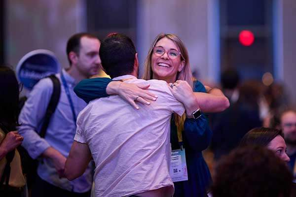 A smiling woman wearing glasses hugs a man at an indoor Gainsight Pulse event. She appears happy and wears a conference badge, with other people visible in the blurred background.