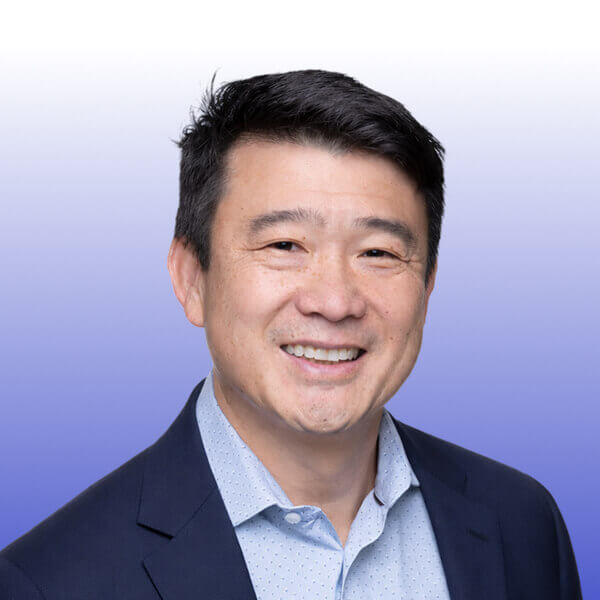 Portrait of Ty Nam, a smiling man with short dark hair wearing a navy blazer and light blue shirt, posing in front of a gradient background that fades from white at the top to purple at the bottom, reminiscent of the signature Gainsight Pulse event style.