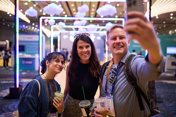 Three people smile and pose for a selfie at an indoor Gainsight Pulse event with colorful neon lights and cloud decorations hanging from the ceiling. Two hold drinks, and they all appear happy and engaged.