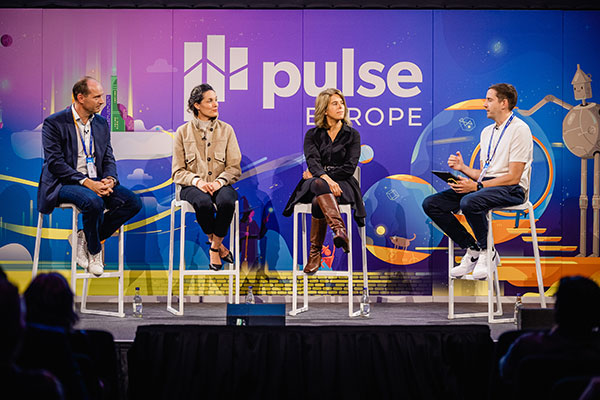 Four people sit on high chairs on stage having a panel discussion at Gainsight Pulse Europe. A colorful backdrop displays the event's name and graphic designs, with the audience visible in the foreground.
