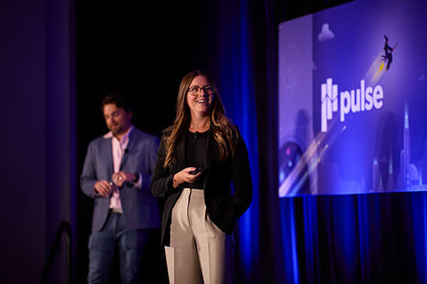 A woman in business attire smiles and presents on stage in front of a large Gainsight Pulse conference screen, while a man in a blazer stands in the background.