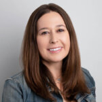 A woman with straight brown hair smiles at the camera. She is wearing a denim jacket over a light top and is posed against a plain light gray background.