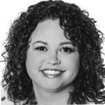 A smiling woman with curly hair, wearing a light-colored top, photographed in black and white against a white background.