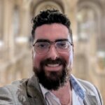 A man with dark hair, a beard, and glasses smiles at the camera. He is wearing a light gray blazer over a white shirt, and the background is a softly blurred, ornate interior with arches.