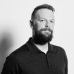 Black and white portrait of a man with short hair and a full beard, wearing a dark polo shirt, looking slightly to the side with a neutral expression against a plain background.