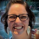 A woman with short brown hair and glasses smiles widely. She is wearing large white earrings and a striped top. The background shows blurred lights and what appears to be water or a waterfront scene.