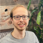 A young man with light skin, short blond hair, glasses, and a trimmed beard smiles at the camera. He is wearing a gray sweater and sits in front of a wooden wall with a green plant beside him.
