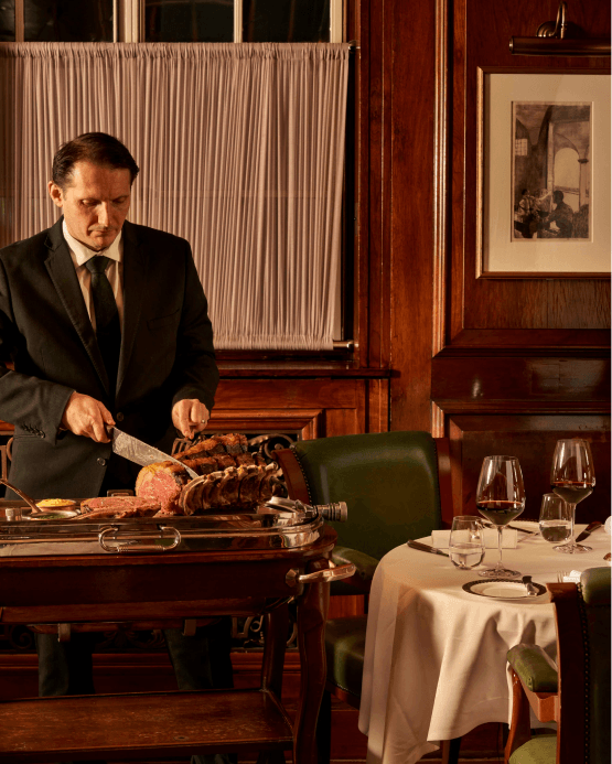 A man in a suit carves meat at a serving cart in an elegant wood-paneled dining room. A set table with wine glasses sits nearby.