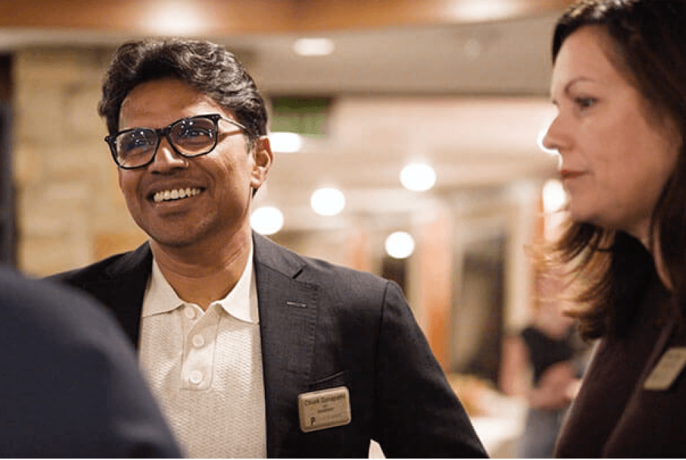 A man in glasses and a dark blazer smiles while talking to a woman in a busy indoor setting. Both are wearing name badges.